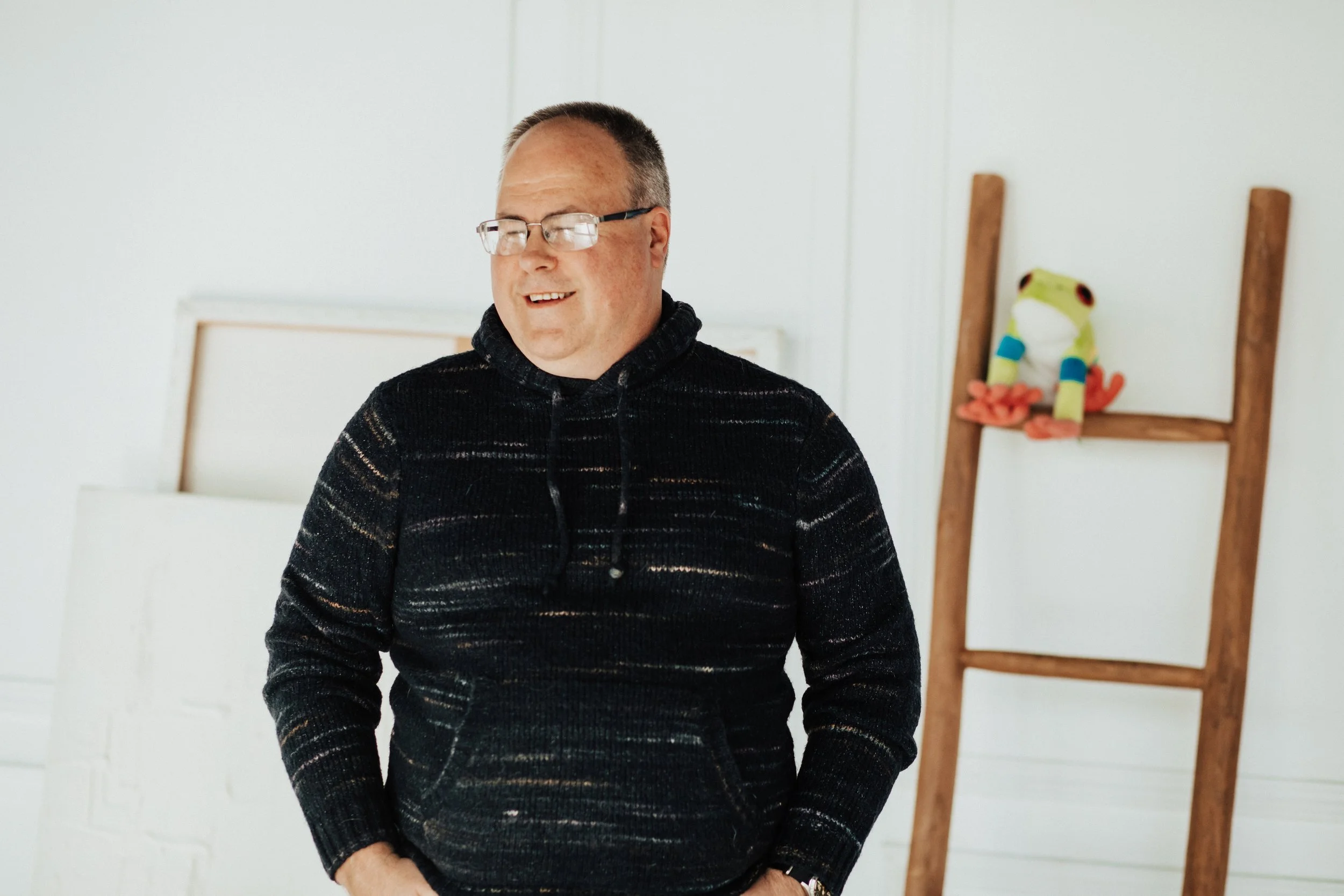 A smiling man with glasses wearing a black patterned hoodie standing indoors near a ladder with a plush frog toy on it.