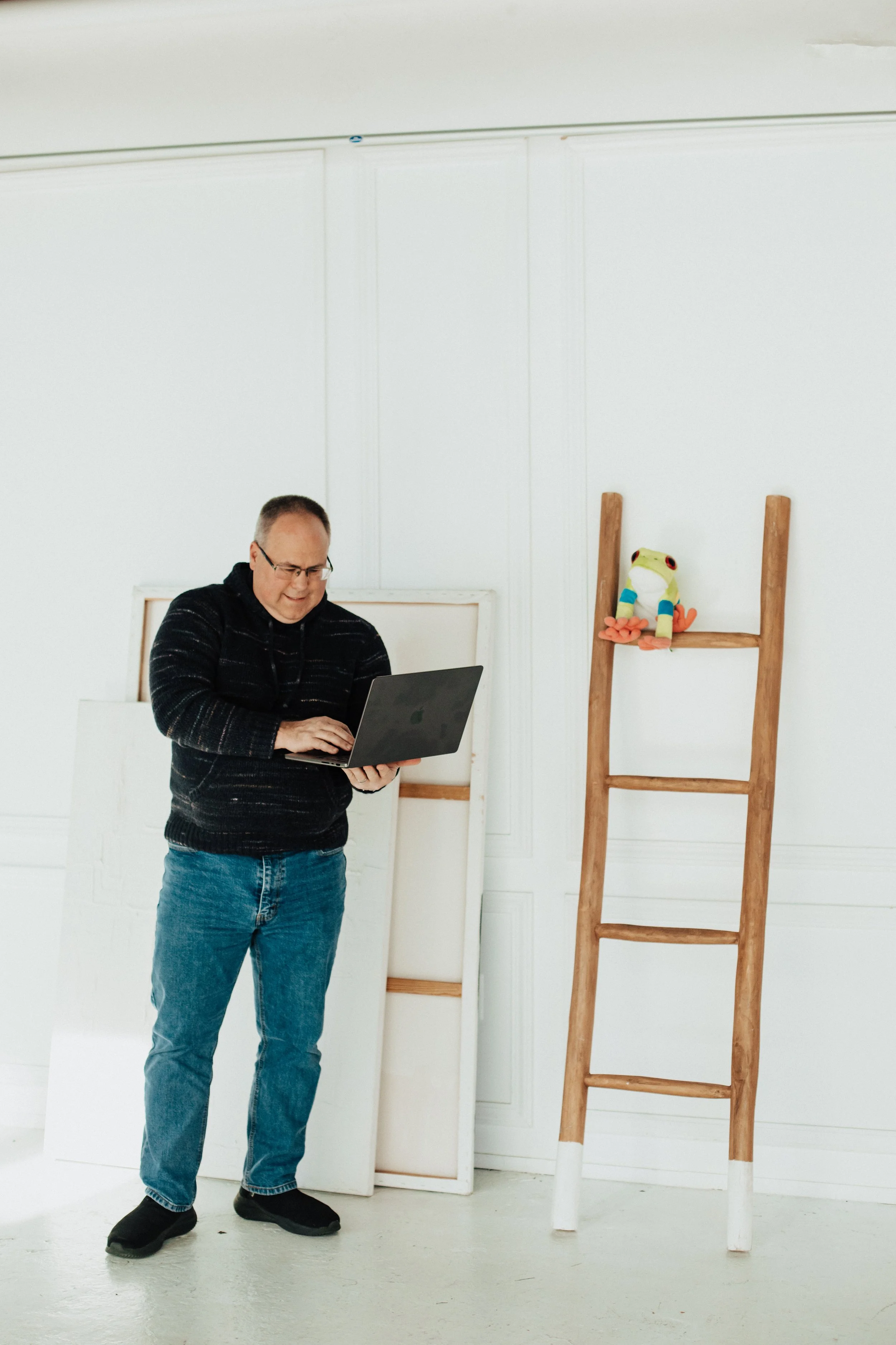 A man stands in a room smiling at a laptop he is holding, with a ladder and picture frames behind him and a stuffed frog on the ladder.