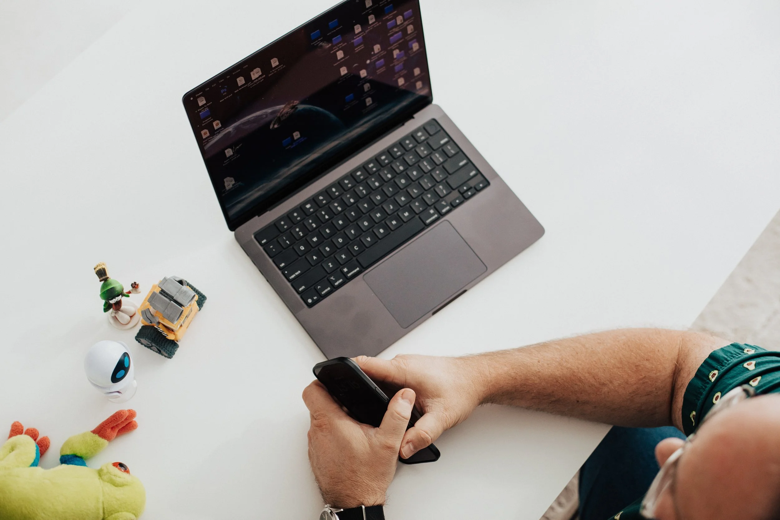 Top view of a person's right arm holding a black remote control, a silver laptop with a space-themed desktop background, and small toys on a white desk, including a yellow toy with red feet, a white robot, a green dinosaur with orange hair, and a small tank.