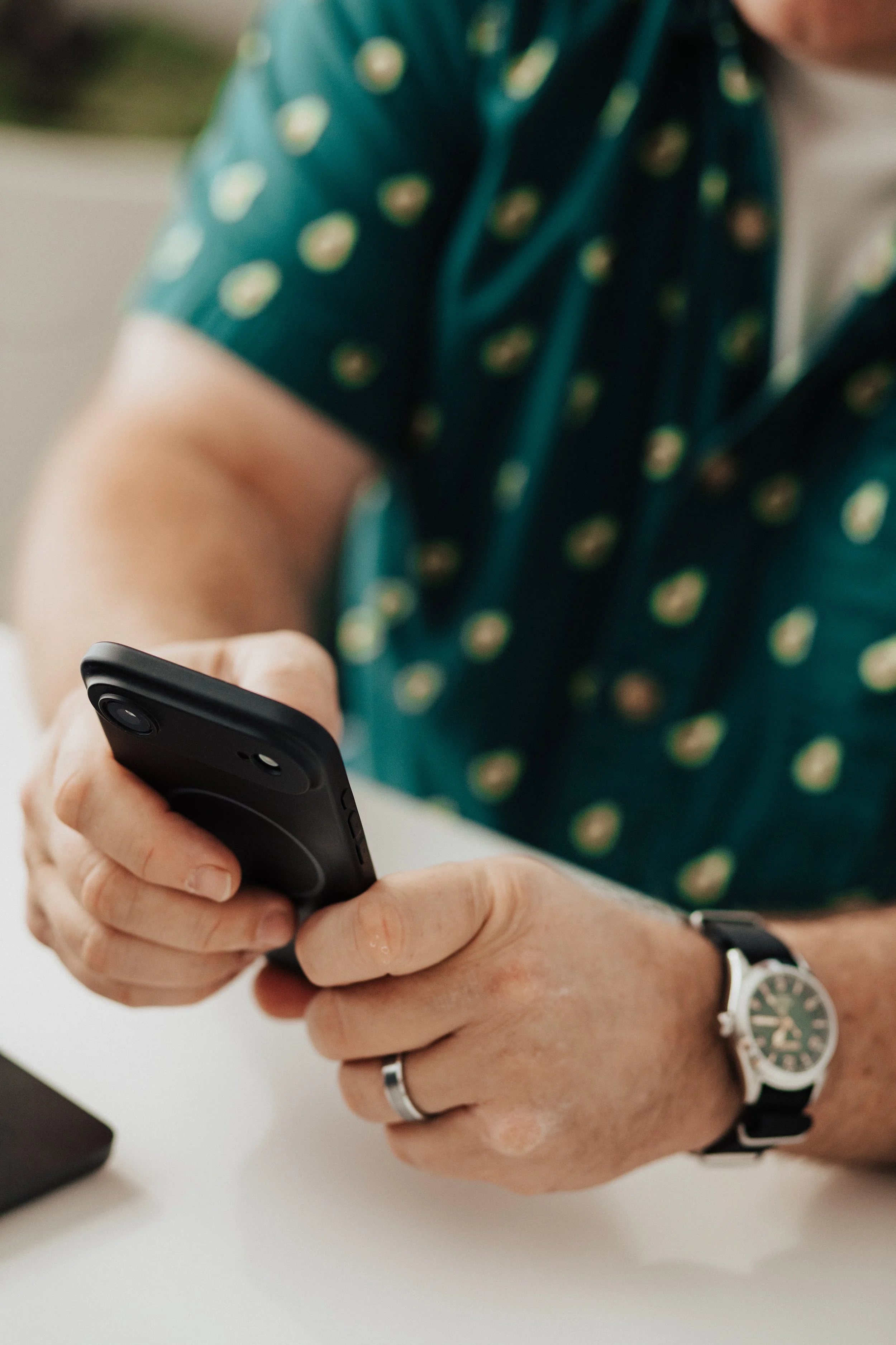 Close-up of a person's hands holding a smartphone, wearing a wedding band on the ring finger, a wristwatch on the left wrist, and a shirt with a pattern of small round figures.