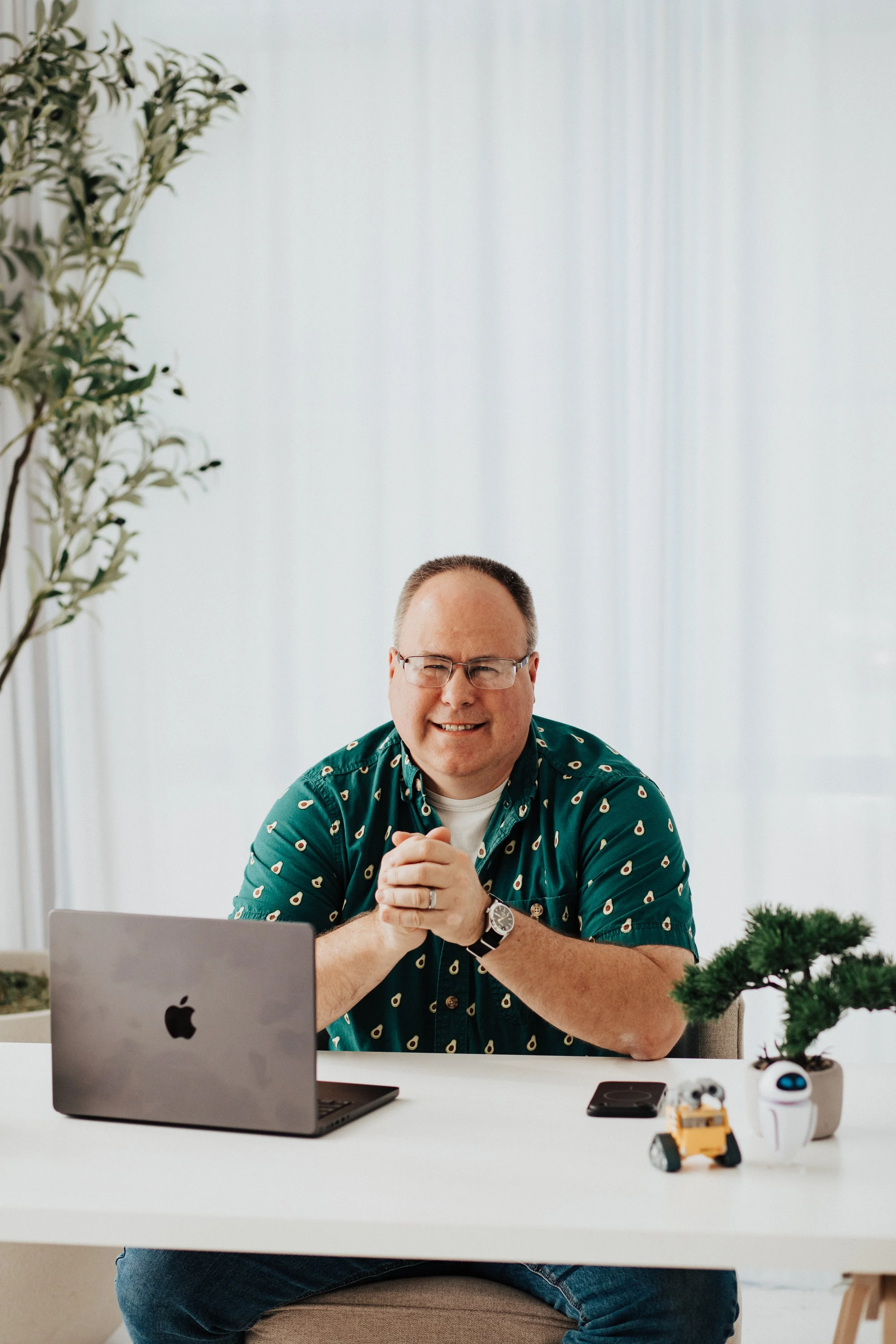 A man with glasses and a patterned teal shirt sits at a white desk with a silver laptop, a small potted plant, a toy robot, and a remote control. He is smiling and clasping his hands together.
