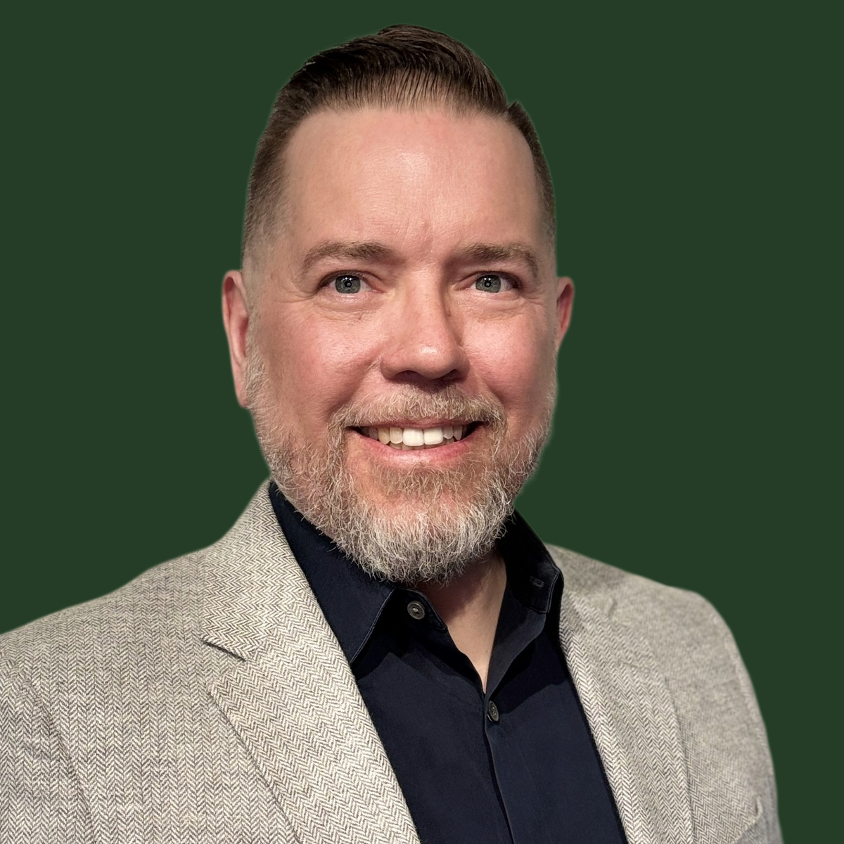 A smiling man with a beard and short hair wearing a light gray blazer and a dark shirt against a dark green background.