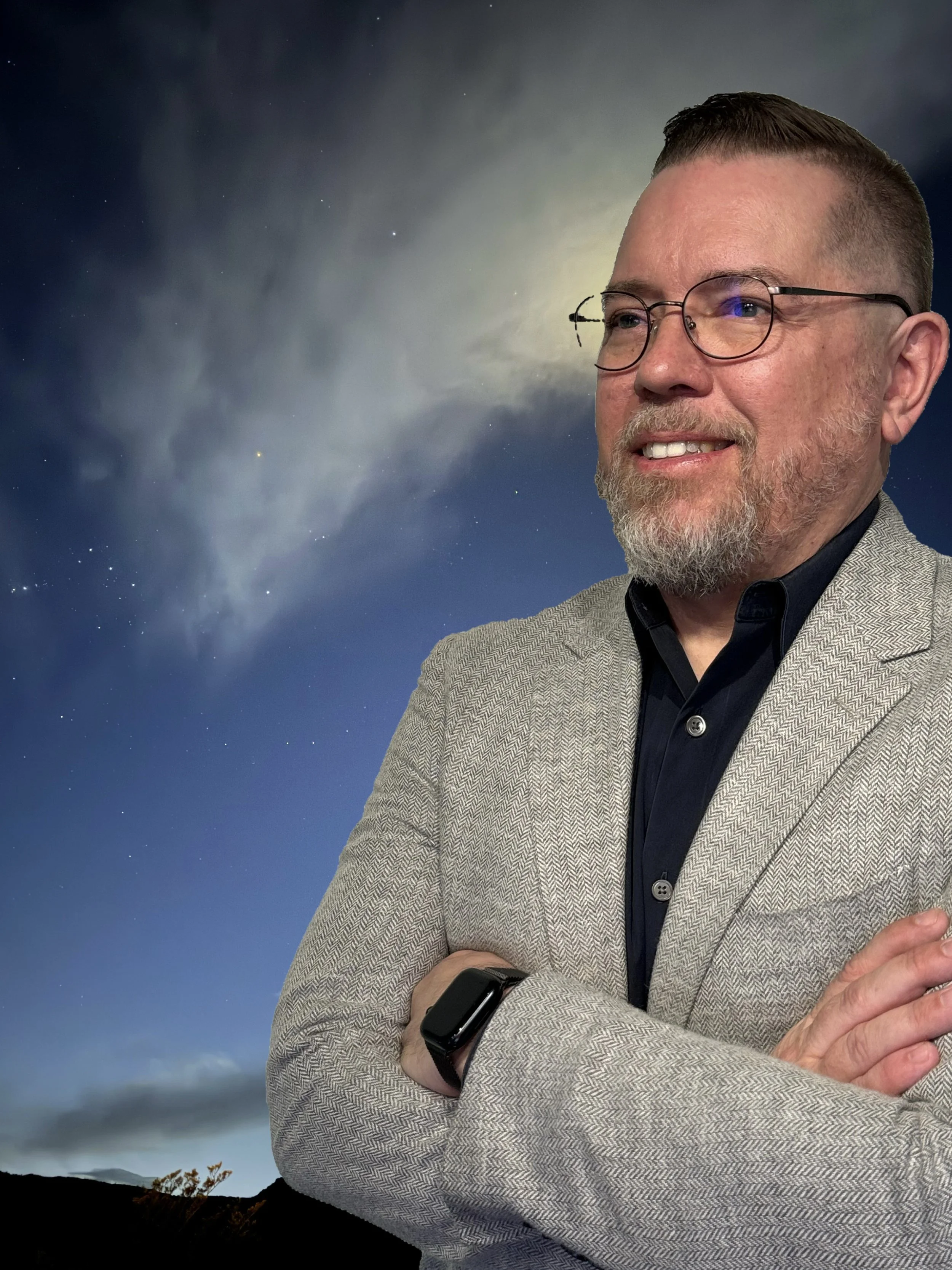 A man in glasses, a gray blazer, and a black shirt stands outdoors with his arms crossed at night. The sky behind him is clear with stars and a bright moon or planet, partially covered by clouds.