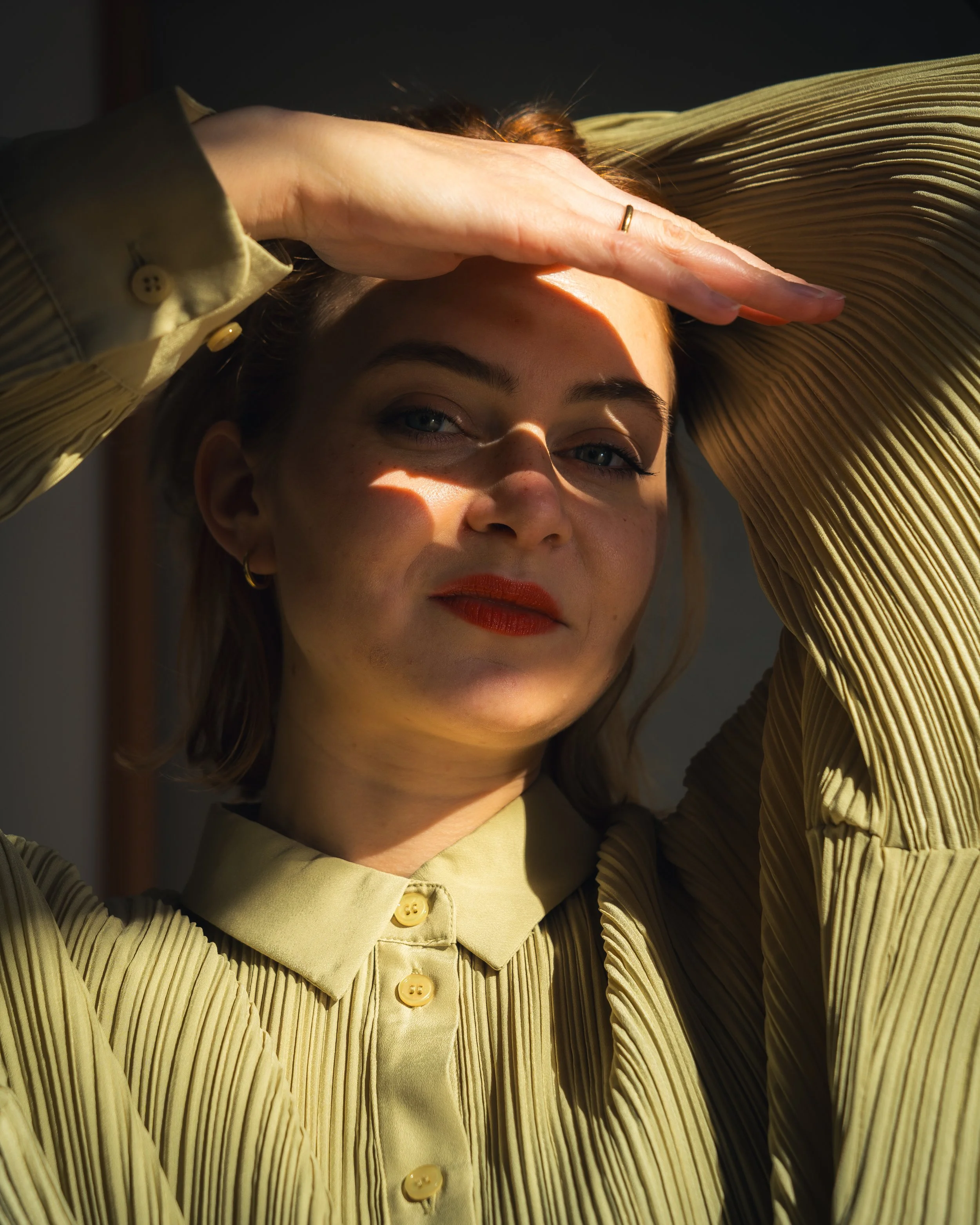 Ein Frau trägt eine gelbe, gerippte Bluse, schaut in die Kamera, hält die Hand über die Stirn, Sonnenlicht scheint auf ihr Gesicht, im Hintergrund ist ein dunkler Raum.