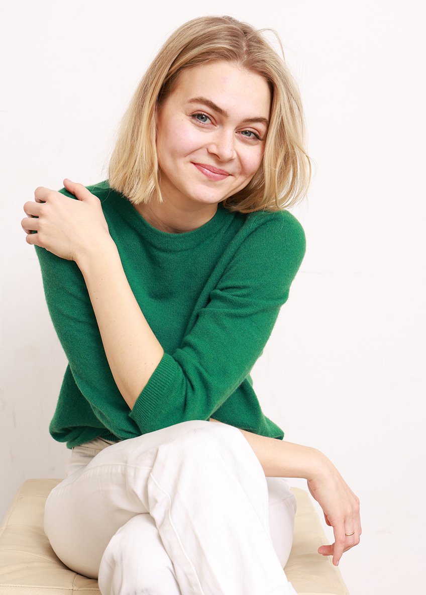 Junge Frau mit blonden Haaren, grüner Pullover und weißen Hosen sitzt lächelnd auf einem Hocker vor weißem Hintergrund.