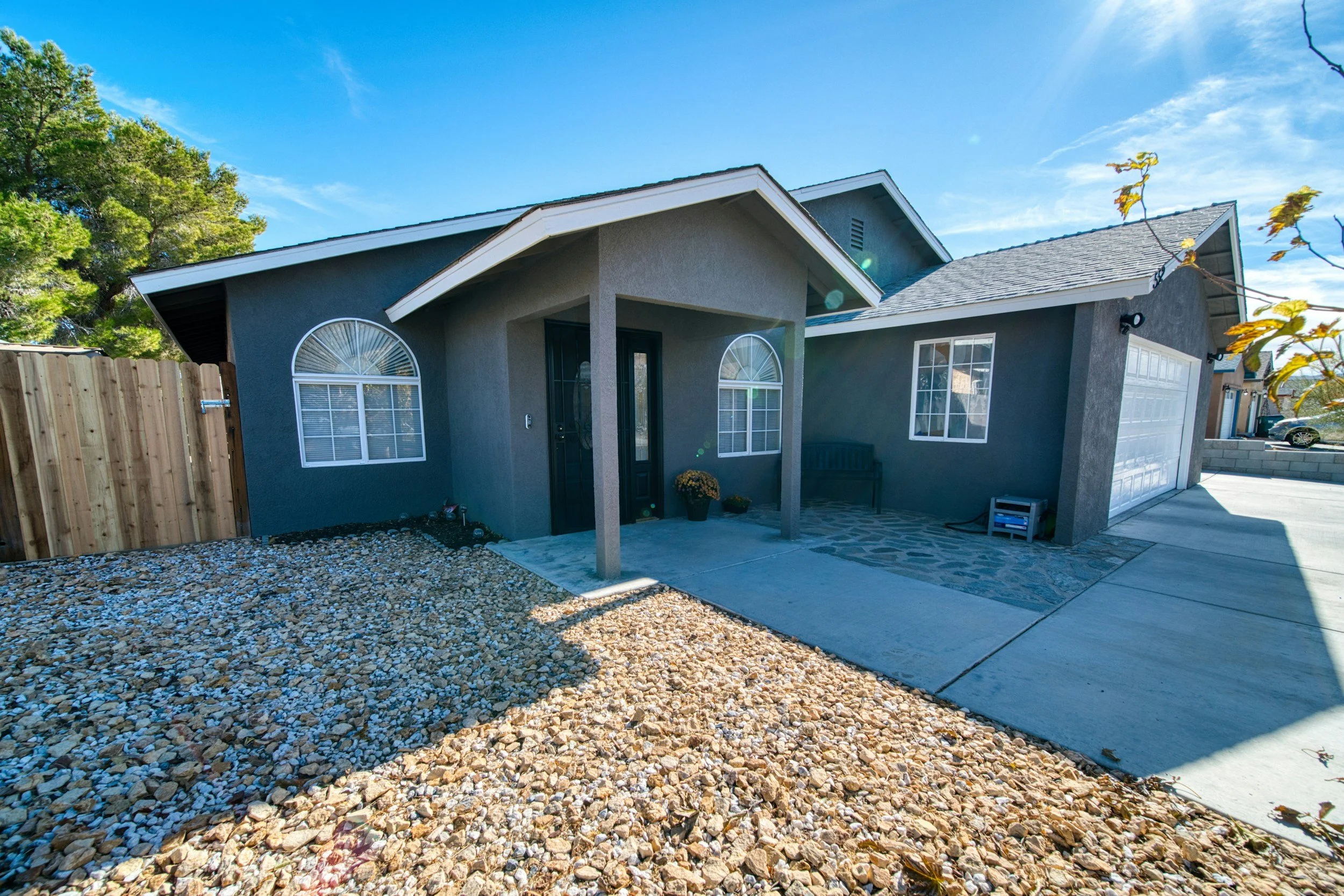 A modern gray house with white trim, a covered front porch, large arched and rectangular windows, and a two-car garage. The front yard has gravel and a concrete driveway under a clear blue sky.