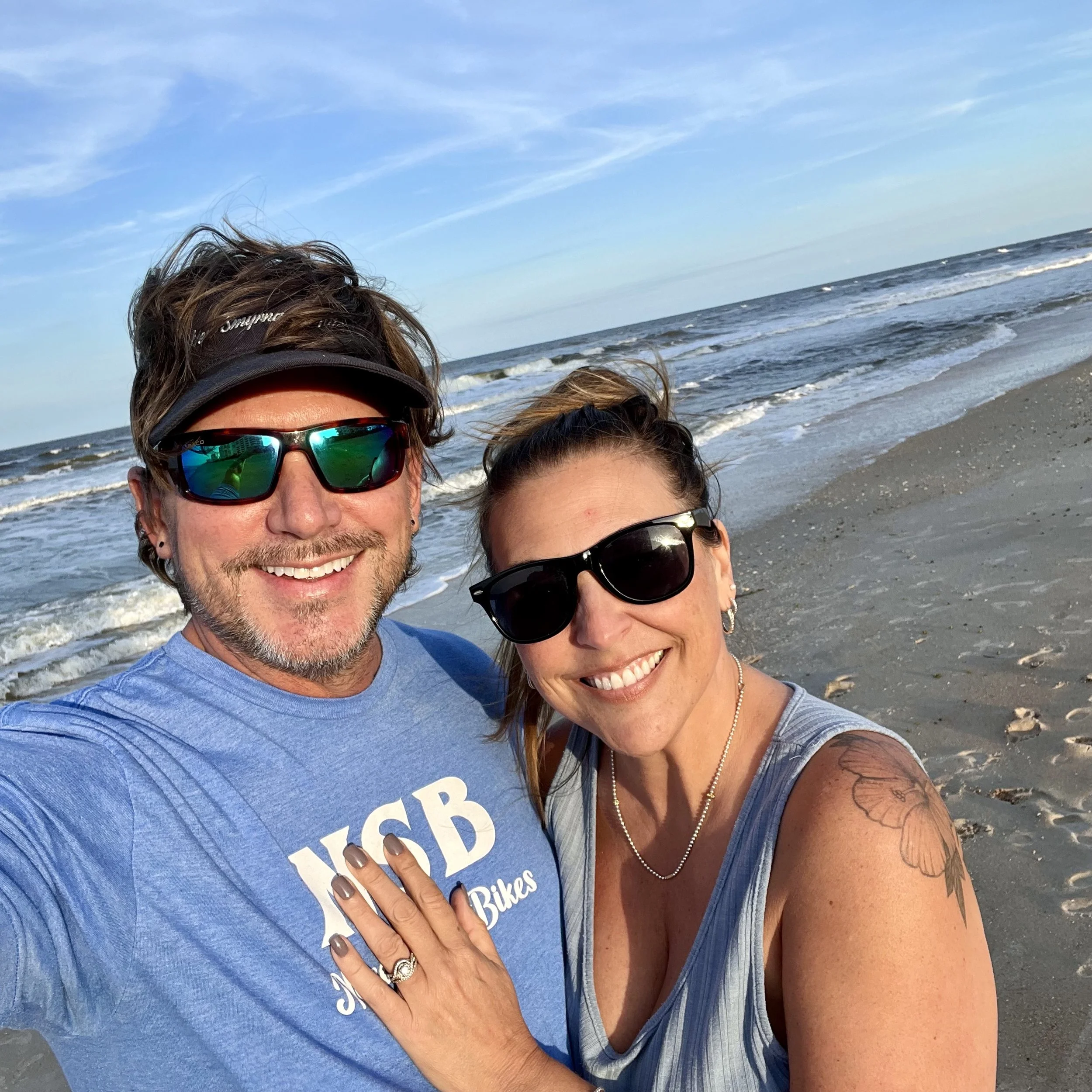 A smiling couple taking a selfie on the beach, with the ocean and a partly cloudy sky in the background. The man is wearing reflective sunglasses, a blue t-shirt, and a visor, while the woman is wearing dark sunglasses, a sleeveless top, and has a tattoo on her shoulder.