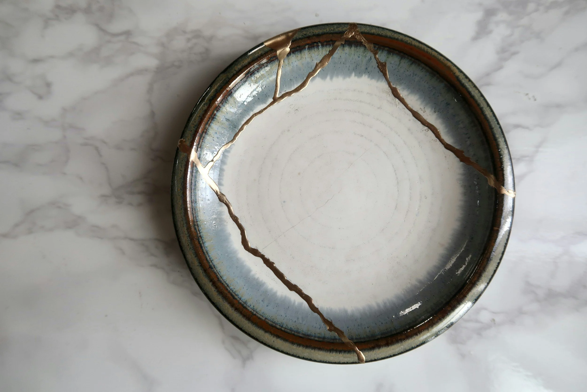 A circular ceramic plate with cracks repaired in the Kintsugi method decorated in blue, brown, and white, placed on a marble surface.