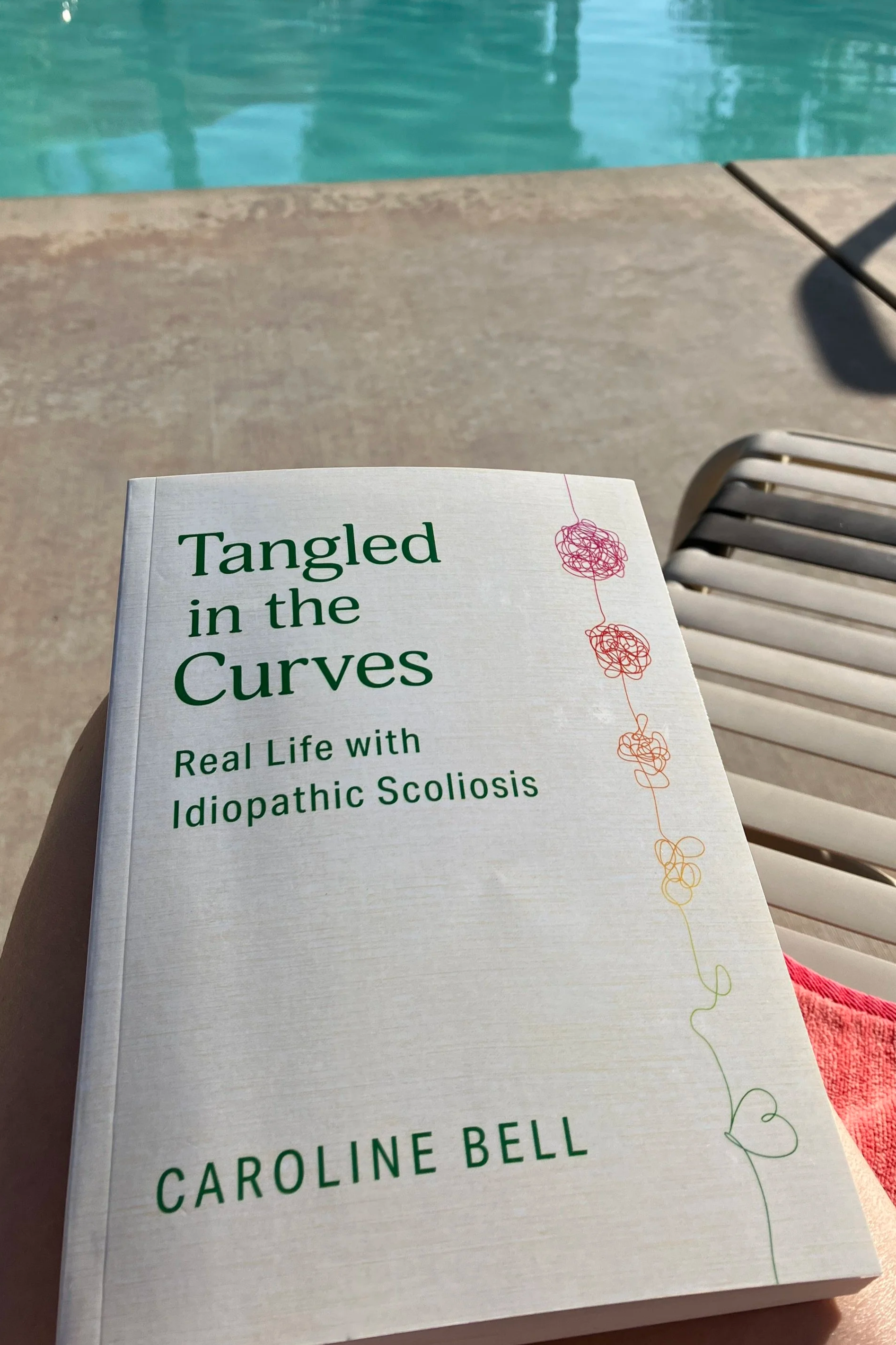 A book titled 'Tangled in the Curves: Real Life with Idiopathic Scoliosis' by Caroline Bell, held in someone's hand near a swimming pool and poolside lounge chair.
