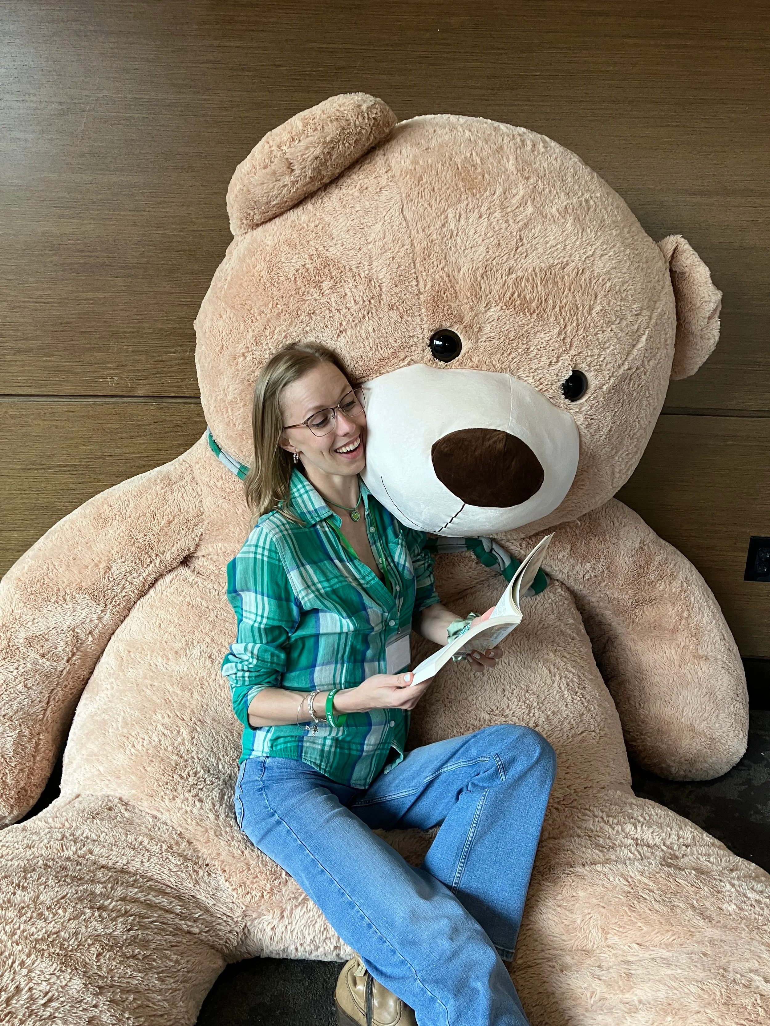 A woman with glasses wearing a green plaid shirt and jeans, sitting next to a giant beige teddy bear holding the book Tangled in the Curves, reading and smiling.