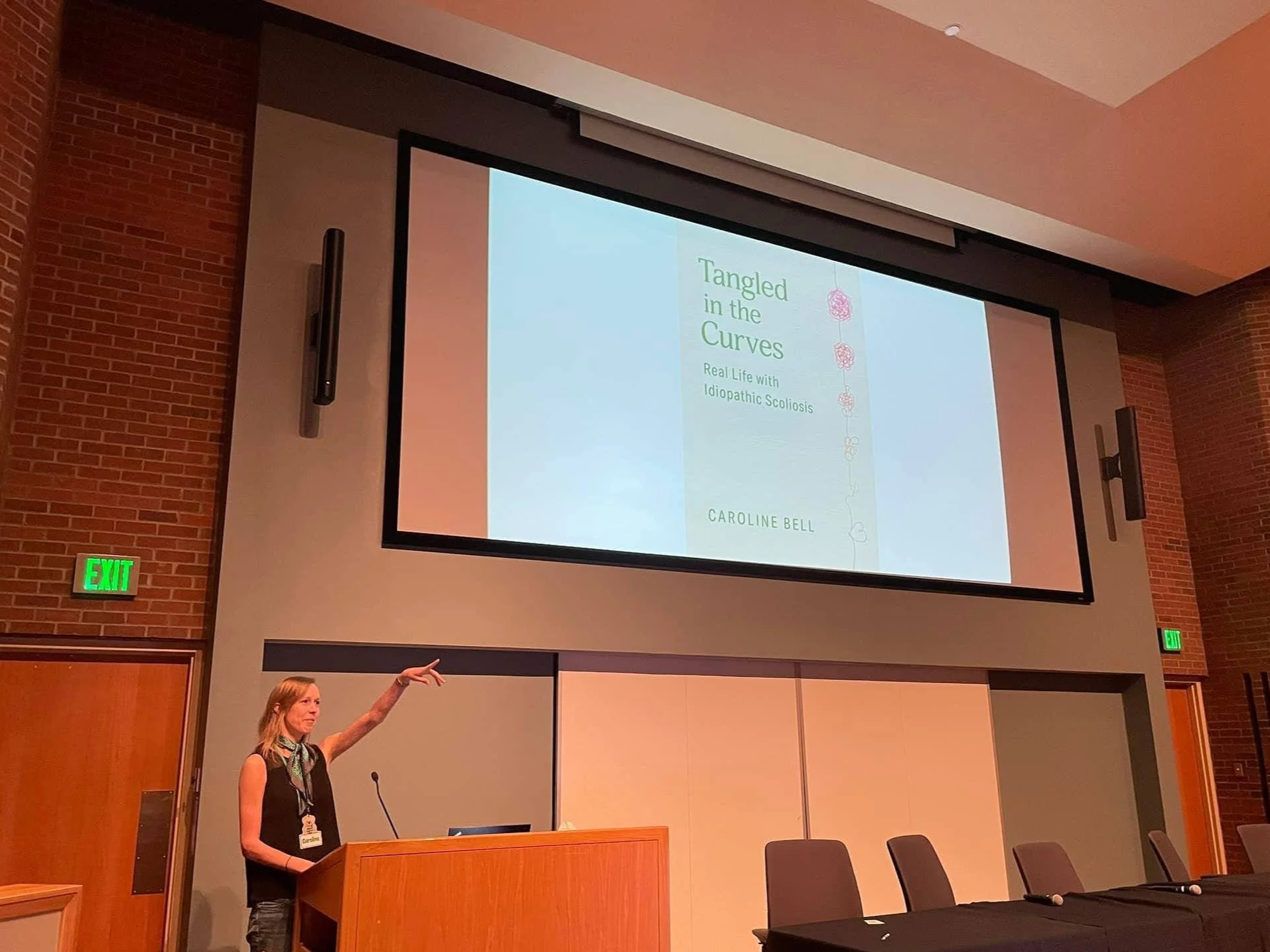 A woman giving a presentation in a conference room with a large screen showing the title 'Tangled in the Curves: Real Life with Idiopathic Scoliosis' by Caroline Bell. The woman is standing behind a podium, pointing to the screen.