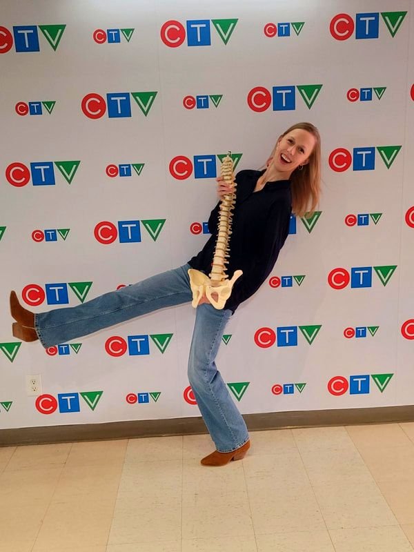 Woman holding a model of a human spine and pelvis, standing in front of a CTV logo backdrop, smiling and balancing with one leg raised.