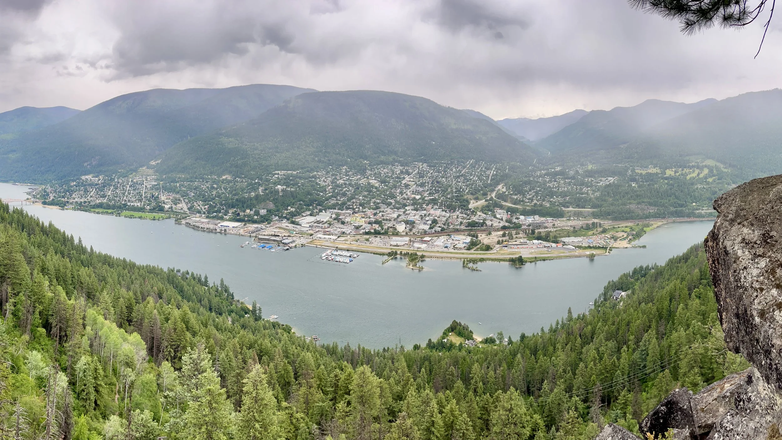 A panoramic view of a city by a river, surrounded by green forests and mountains under a cloudy sky.