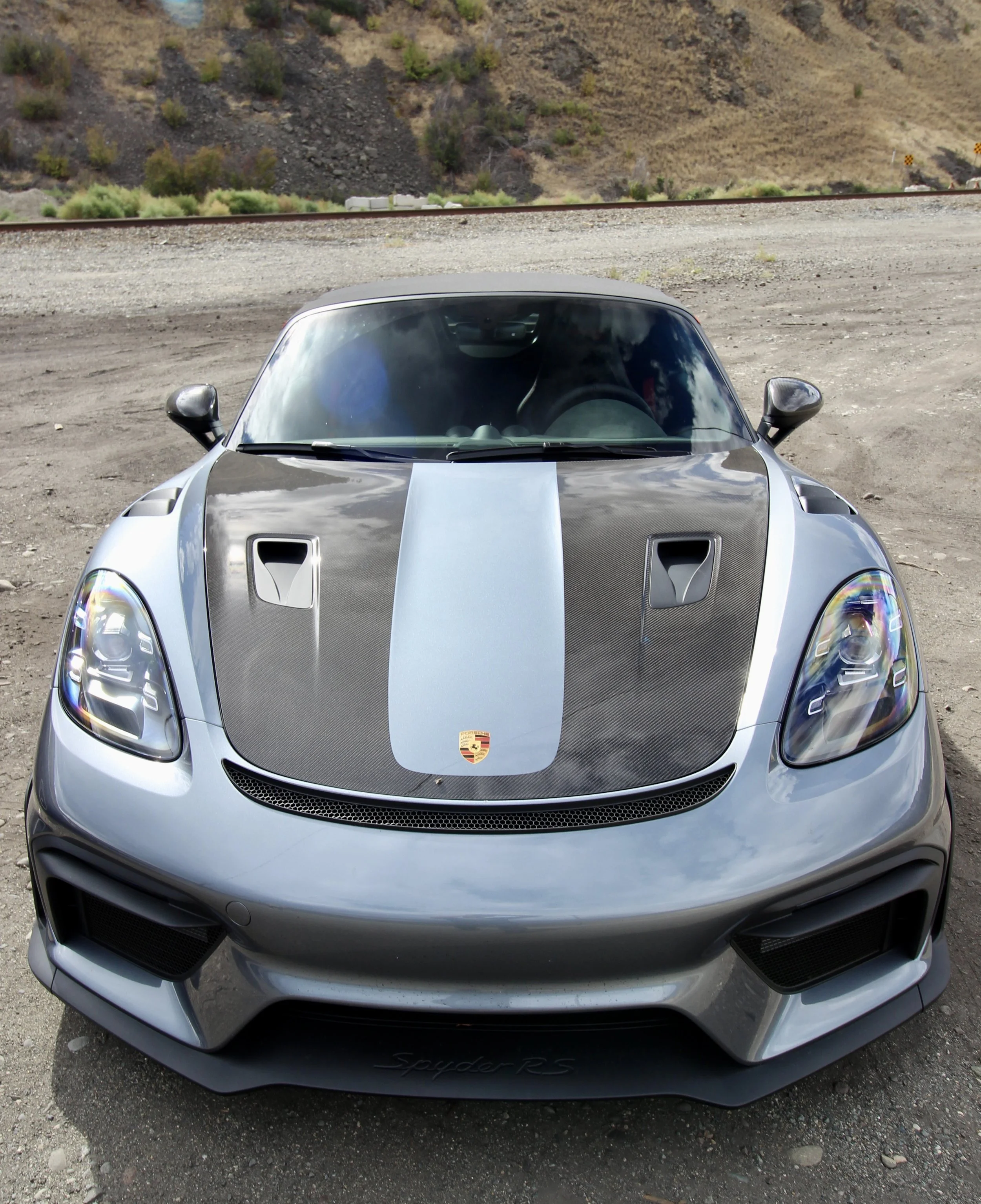 Front view of a silver Porsche sports car with a black and blue racing stripe on the hood, set against a desert landscape with hills and train tracks in the background.