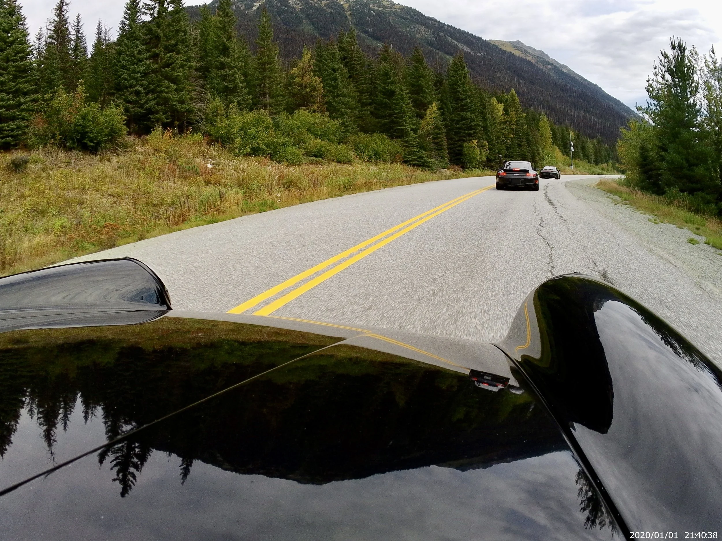 View from a car driving on a mountain road with a double yellow line. The car has a black hood and is surrounded by tall pine trees and mountains in the background. Several other cars are on the road ahead.