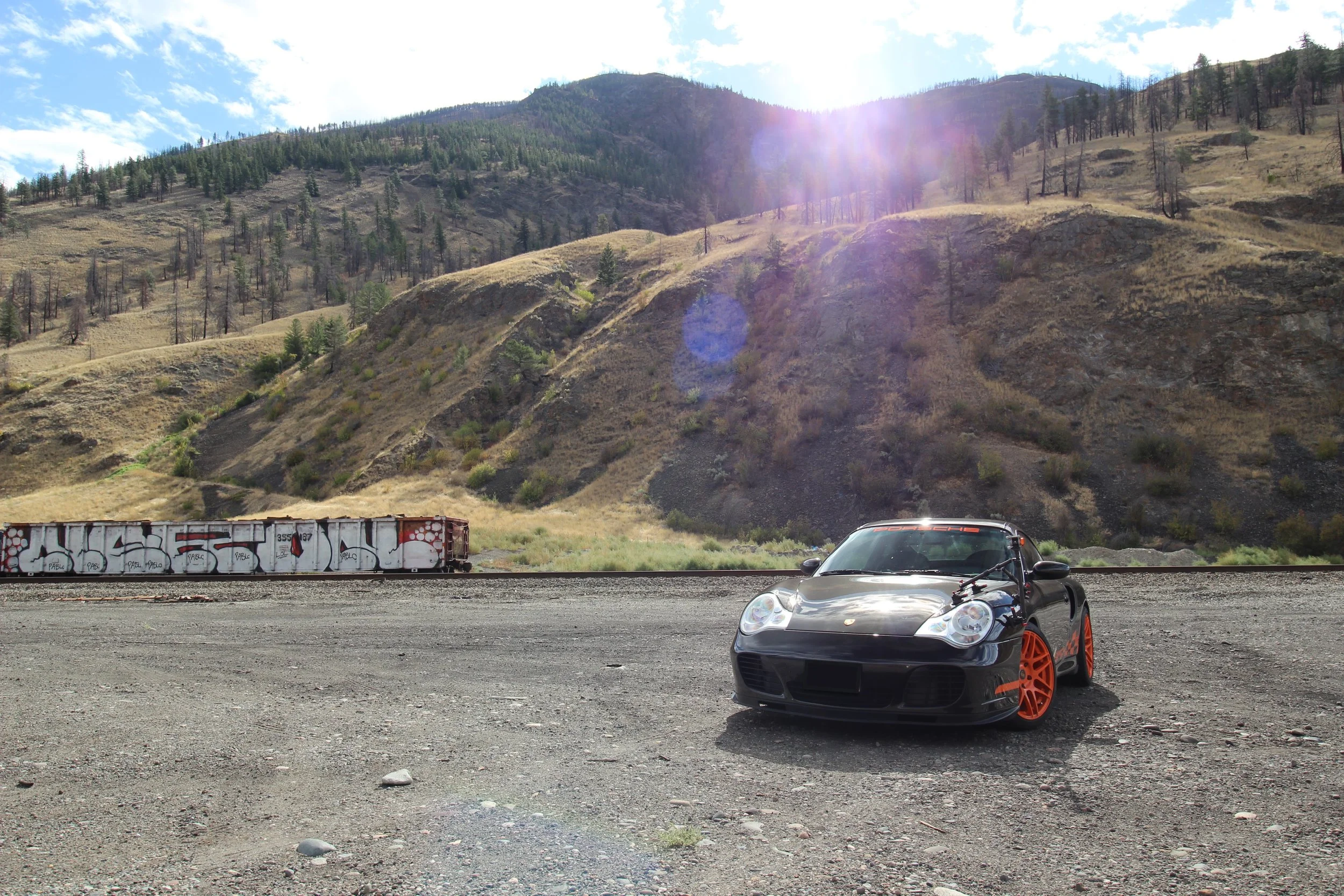 A black Porsche sports car with orange wheels parked on a dirt area with mountain scenery and a train car in the background.