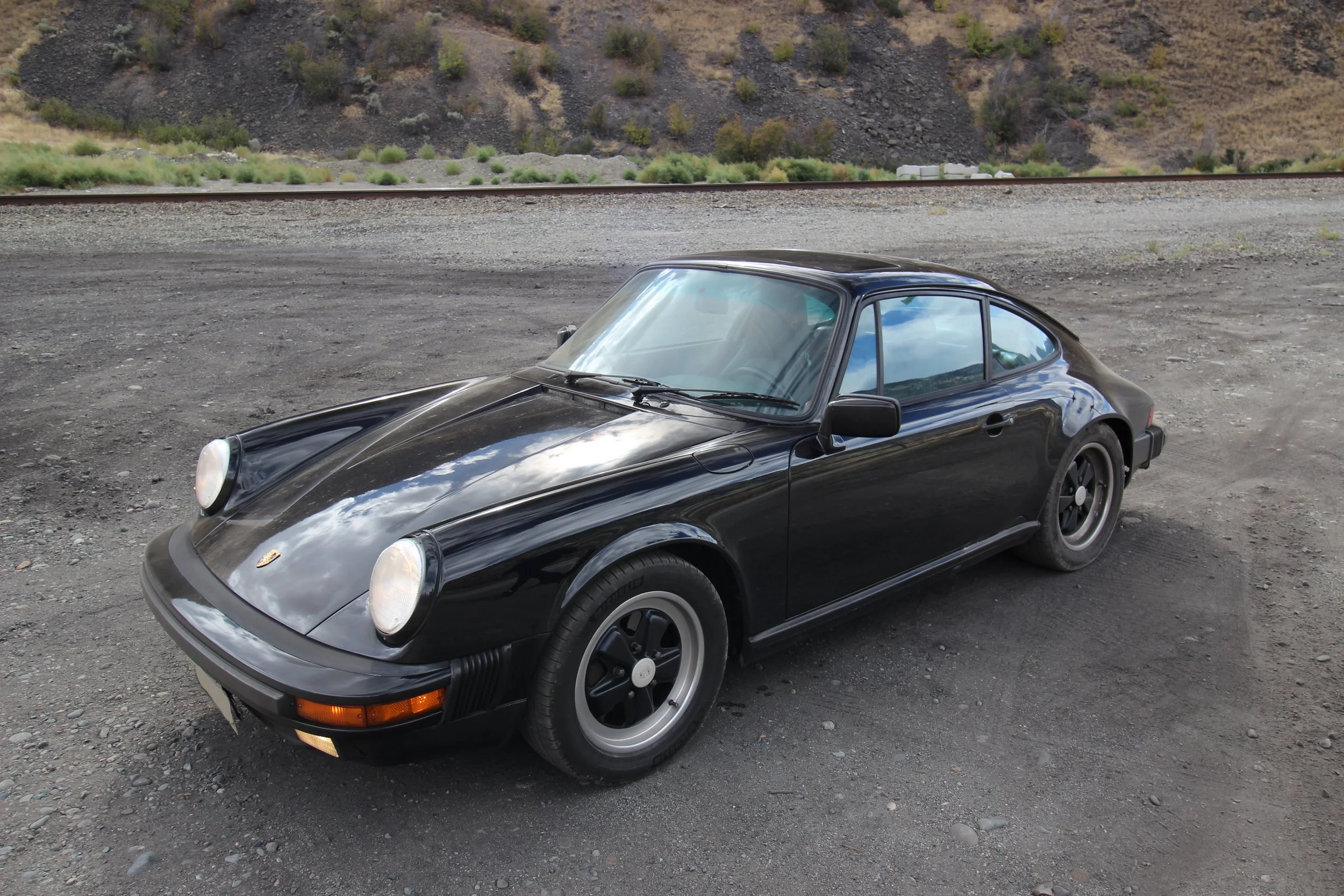A black vintage Porsche 911 sports car parked on a gravel lot with a train track and hillside in the background.