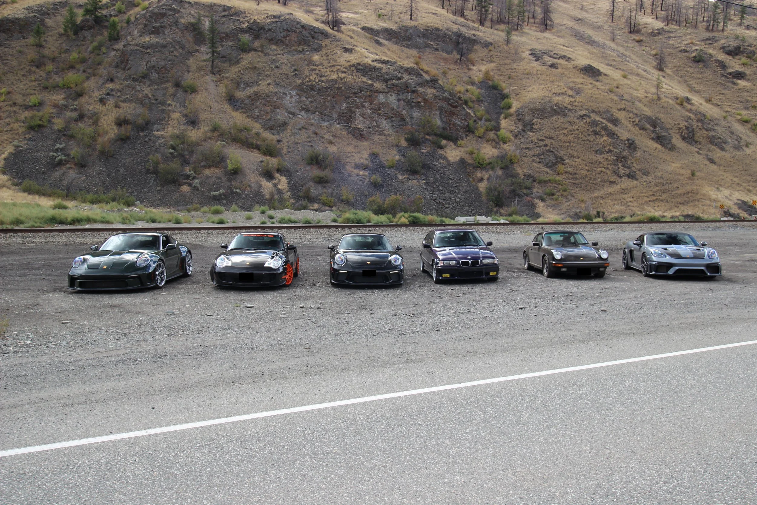 Six high-performance sports cars parked on the side of a road with mountainous terrain in the background.