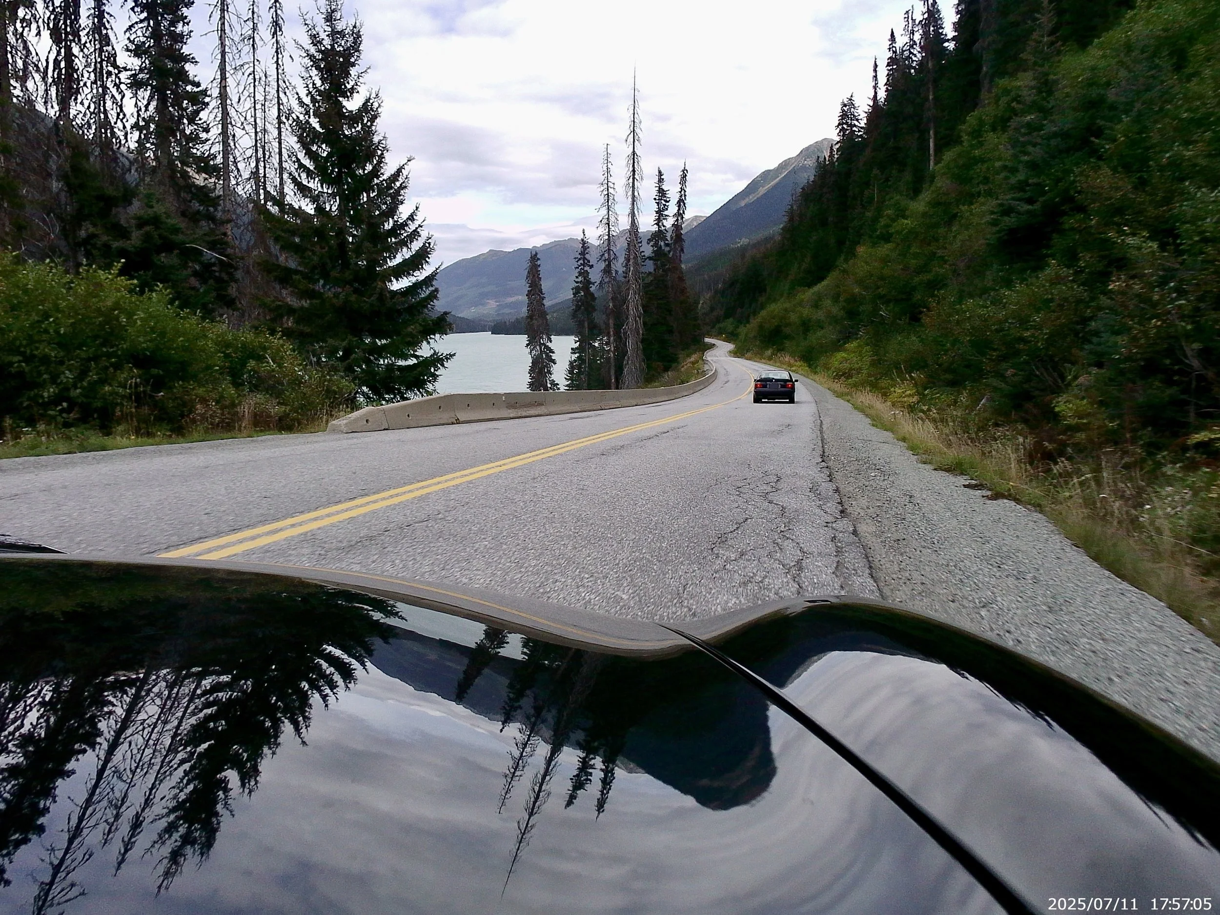 View from inside a vehicle driving on a winding mountain road next to a lake, surrounded by tall pine trees and mountains under a cloudy sky.