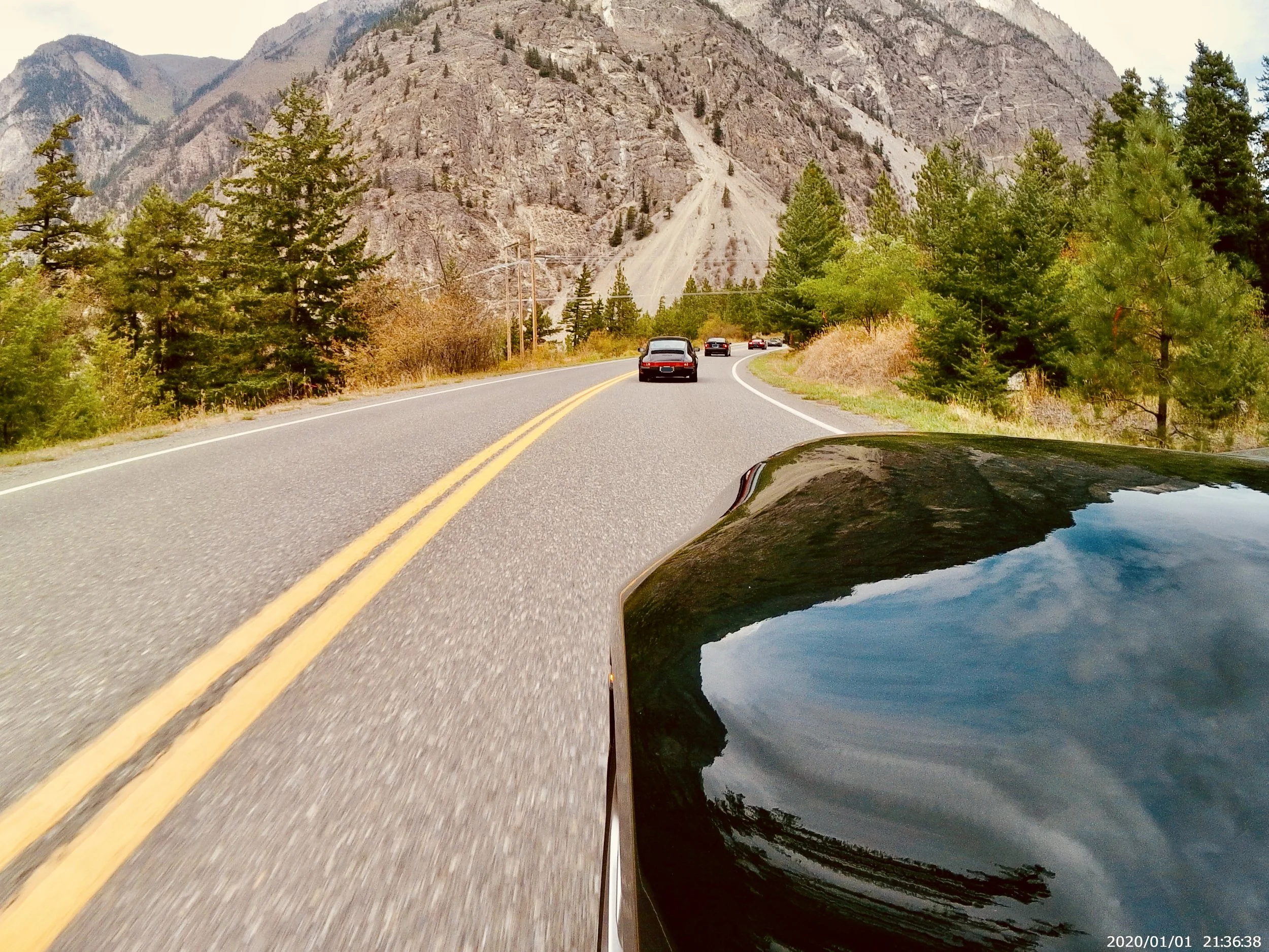 A mountain road with several cars driving along it, surrounded by pine trees and rugged mountains in the background, taken from a moving vehicle.