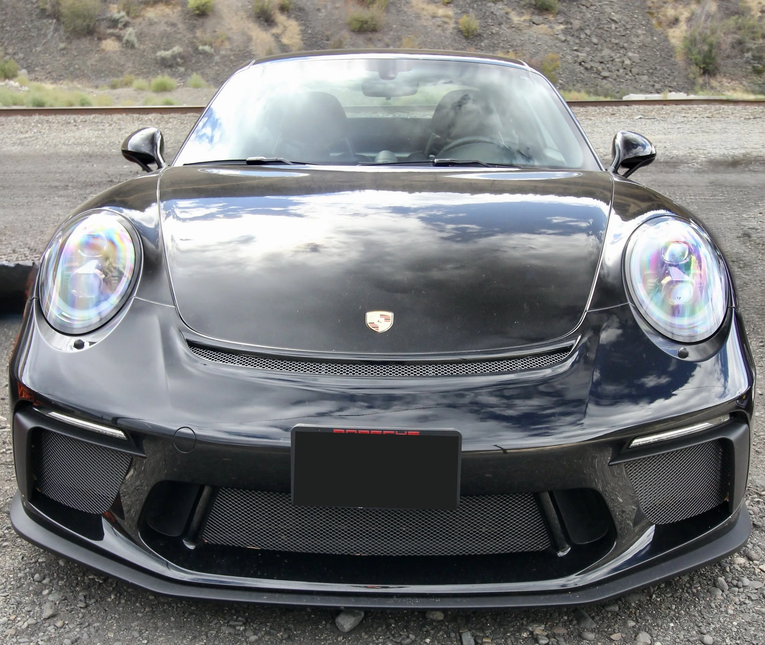 Front view of a black Porsche sports car parked outdoors, with reflections of clouds and sky on its hood and windshield.