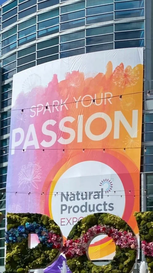A colorful vertical banner on a building promoting a Natural Products Expo with the message 'Spark Your Passion'. In front, there's a floral-themed decorative display shaped like the numbers 2024.