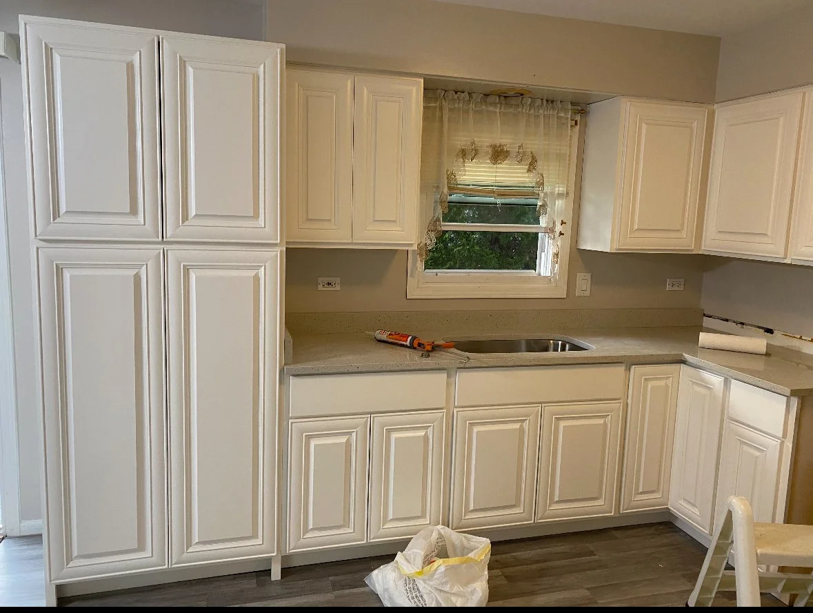 White kitchen cabinets with a window above the sink, beige countertop, and a workspace with tools. A plastic bag is on the floor near a chair.