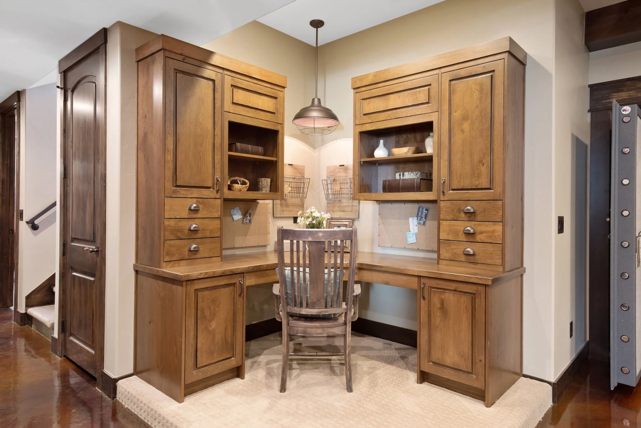Wooden corner desk with matching cabinets and open shelves, a chair, and a hanging pendant light, with decorative items and a flower arrangement on the desk.