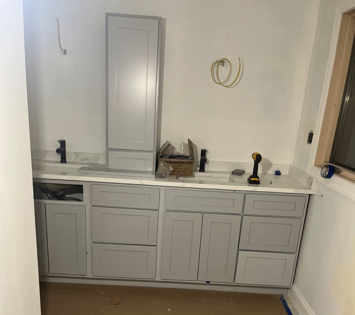 Bathroom vanity under construction with cabinet doors installed, a countertop, and tools such as a drill on the surface; the wall has electrical wiring and piping for a sink.