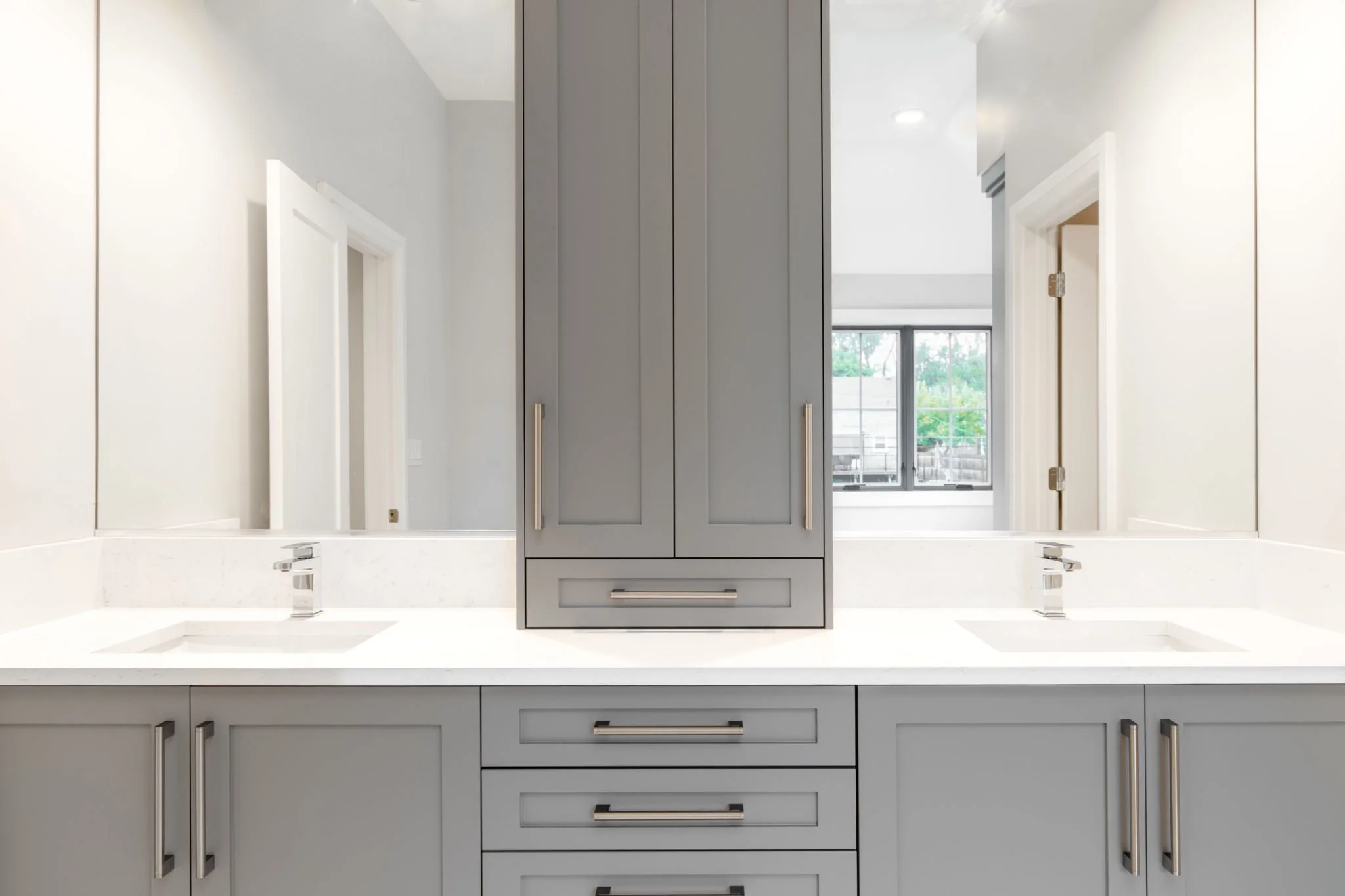 Bathroom vanity with two mirrors, two sinks, and gray cabinets with chrome handles, reflecting a window and a door.