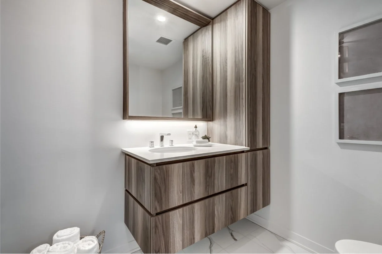Modern bathroom vanity with a large mirror, wooden cabinetry, and a white countertop with a sink and soap dispenser. White towels are stacked in a basket on the floor.