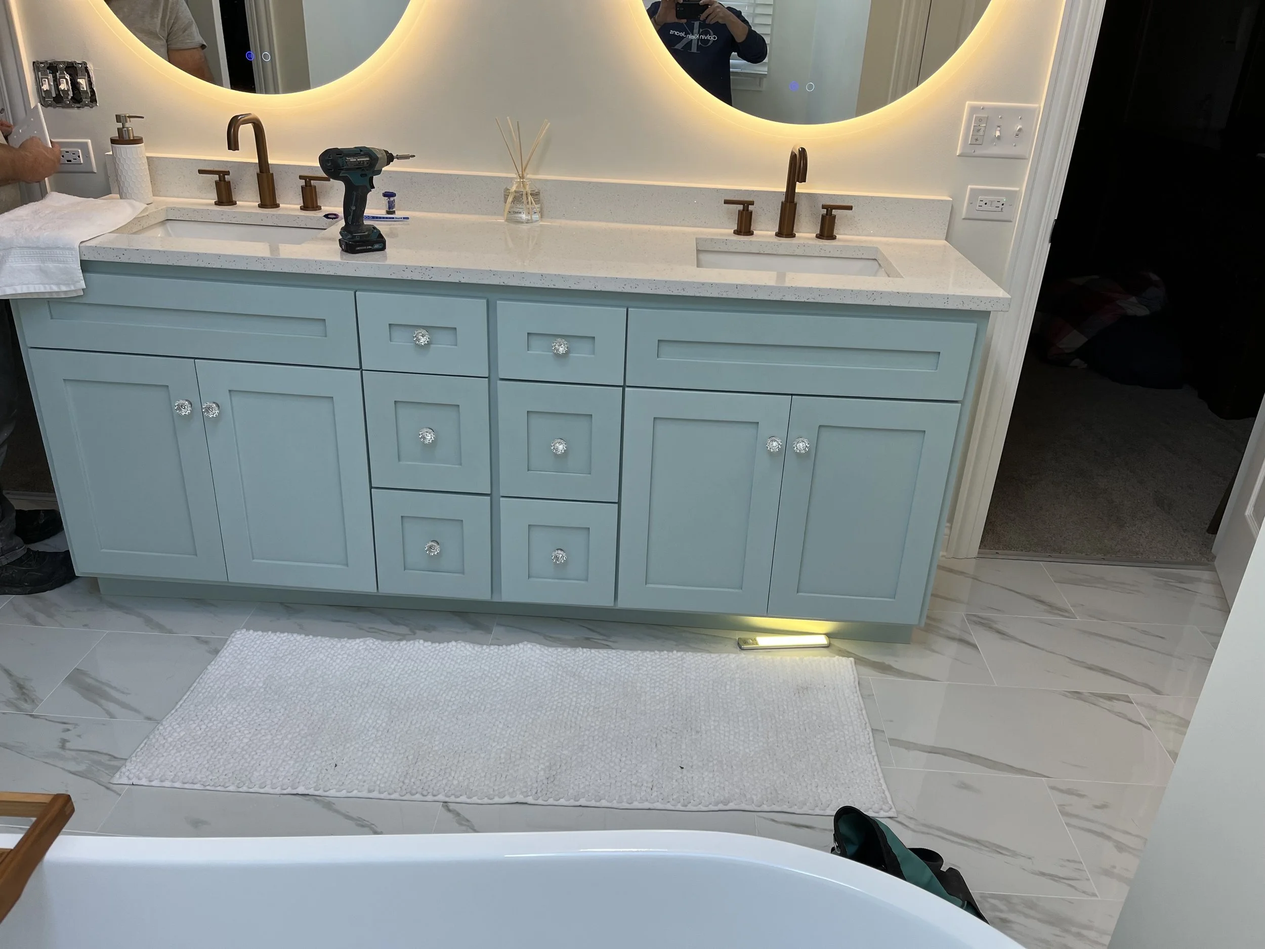 A bathroom vanity with a light blue cabinet, a white countertop, and two round mirrors with backlighting. There is a drill, a soap dispenser, and a reed diffuser on the counter. The floor has marble tiles and a white rug.