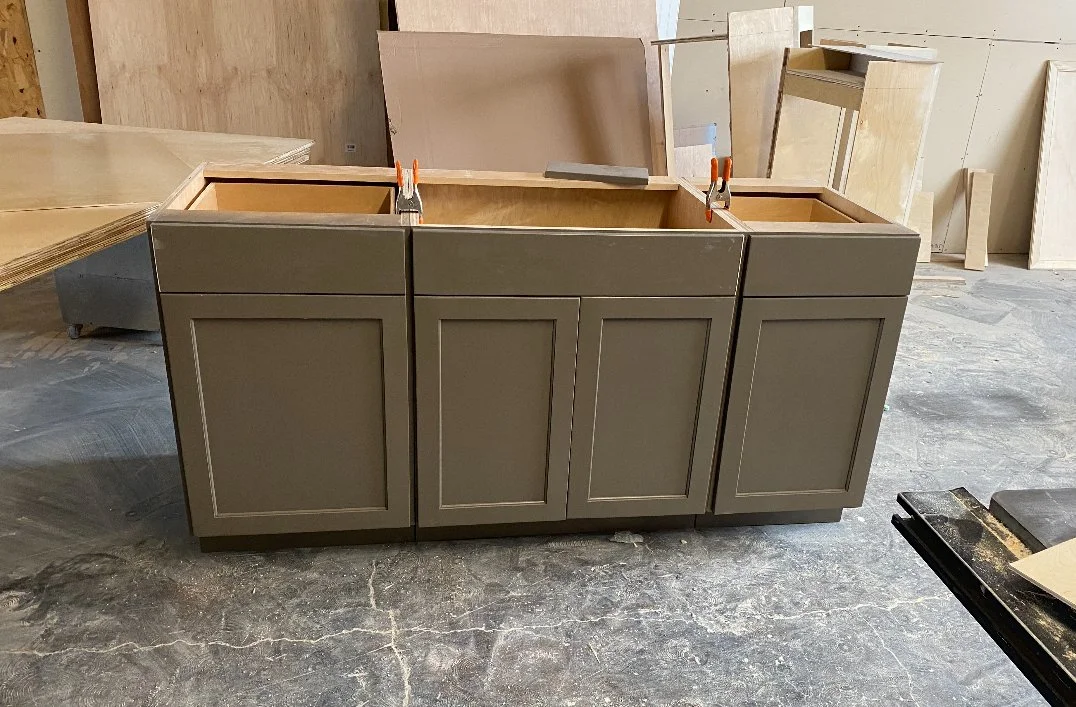 A partially assembled kitchen cabinet with a taupe-colored finish, three doors, and clamps at the top, in a woodworking shop with various wood pieces and panels in the background.
