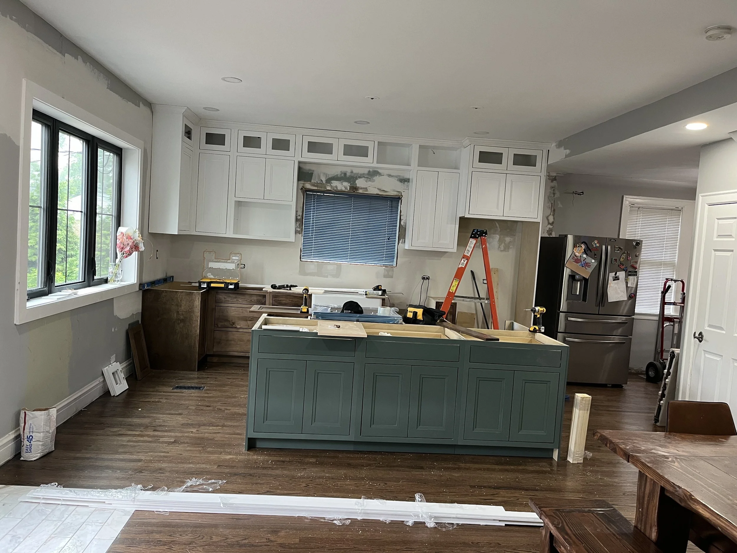 Kitchen under renovation with cabinets being installed, an orange ladder, construction tools, a refrigerator with magnets, and construction materials on the floor.