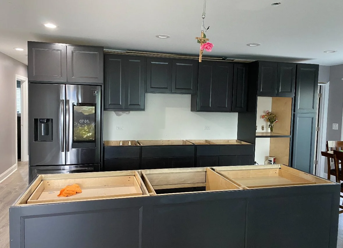 Kitchen under renovation with black cabinets, a stainless steel refrigerator, and a kitchen island frame. There is a small flower arrangement on a built-in shelf on the right wall.