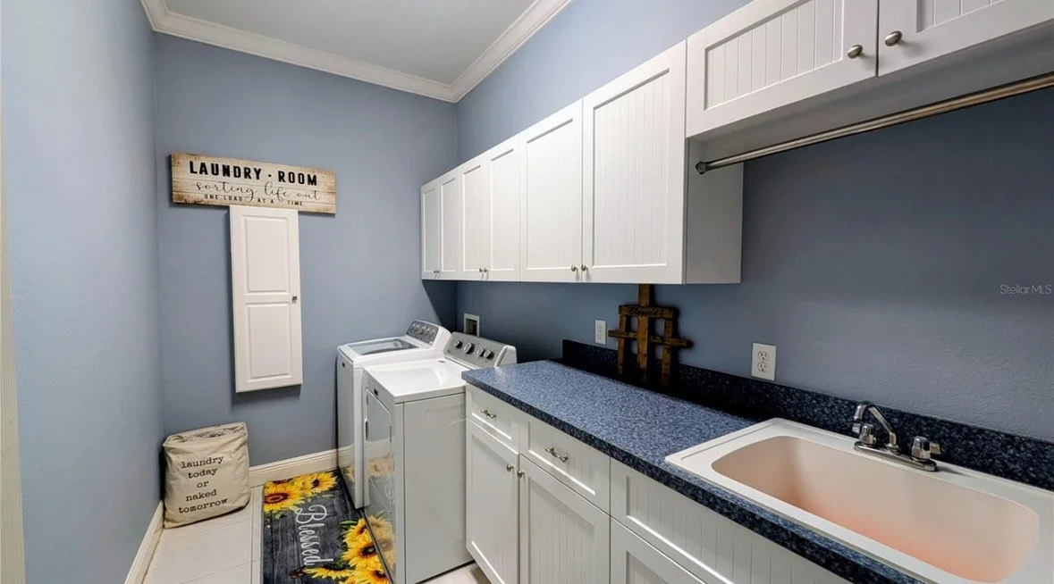 Laundry room with white cabinets, a blue countertop, a beige sink, and a washing machine, with wall decor including a sign and a fabric bag.