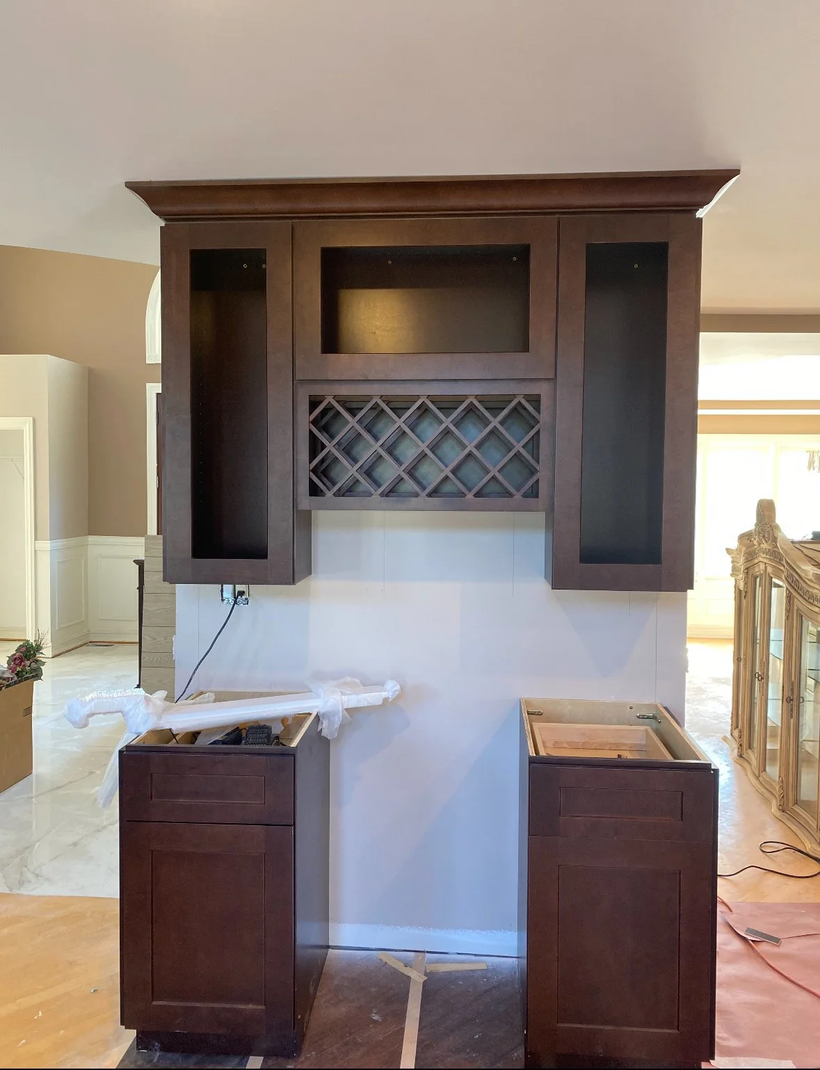 Dark brown kitchen cabinet with open shelves and wine rack, with two empty base cabinets, one with a white wrapping, in a room with beige walls and marble flooring.