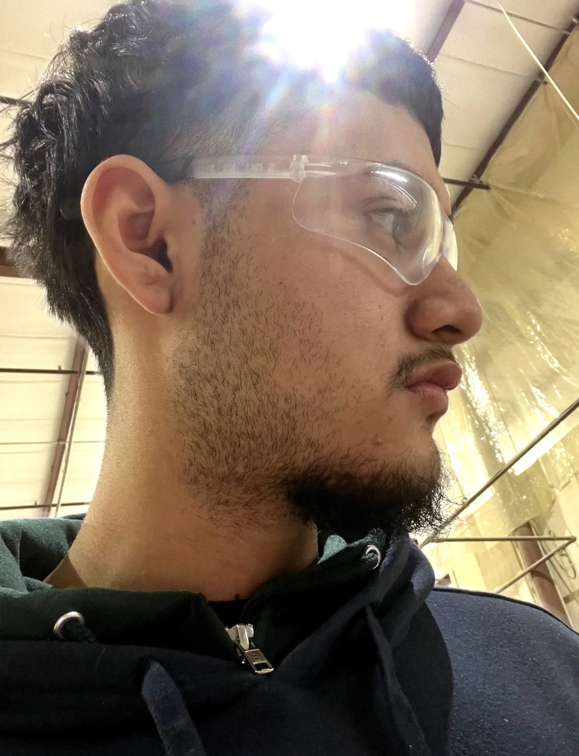 Profile of a young man with dark hair, facial stubble, wearing safety goggles, inside a brightly lit industrial or warehouse setting with a yellowish plastic ceiling and metal supports.