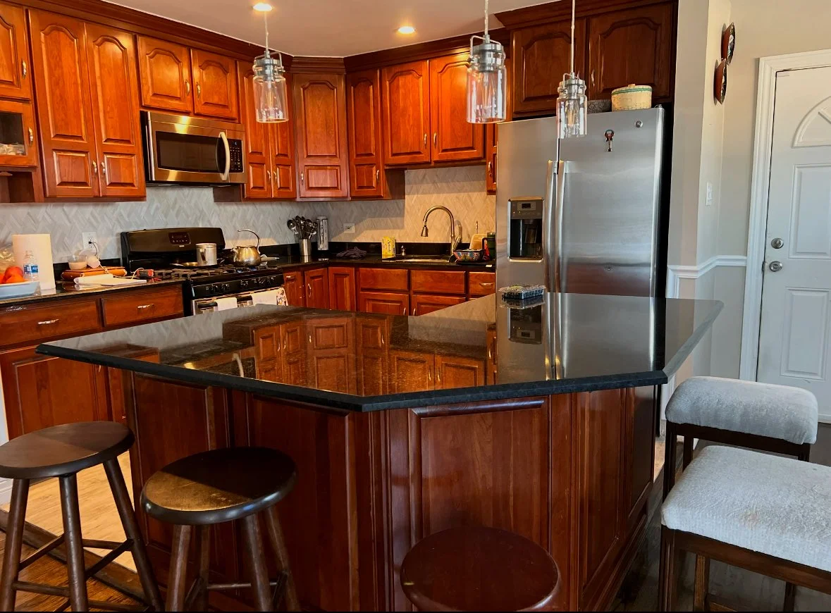 Kitchen with wooden cabinets, black countertop, stainless steel appliances including a microwave and refrigerator, a kitchen island with bar stools, hanging pendant lights, and a white door.