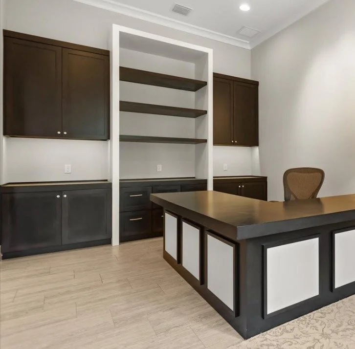 An empty office or conference room with dark wood cabinets and a built-in bookshelf, a wooden desk with a black frame, and a brown office chair.