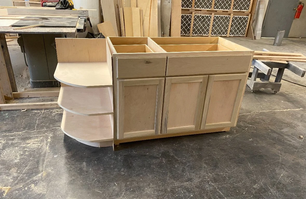 unfinished wooden kitchen cabinet with open shelving and closed cabinet doors in a woodworking shop.