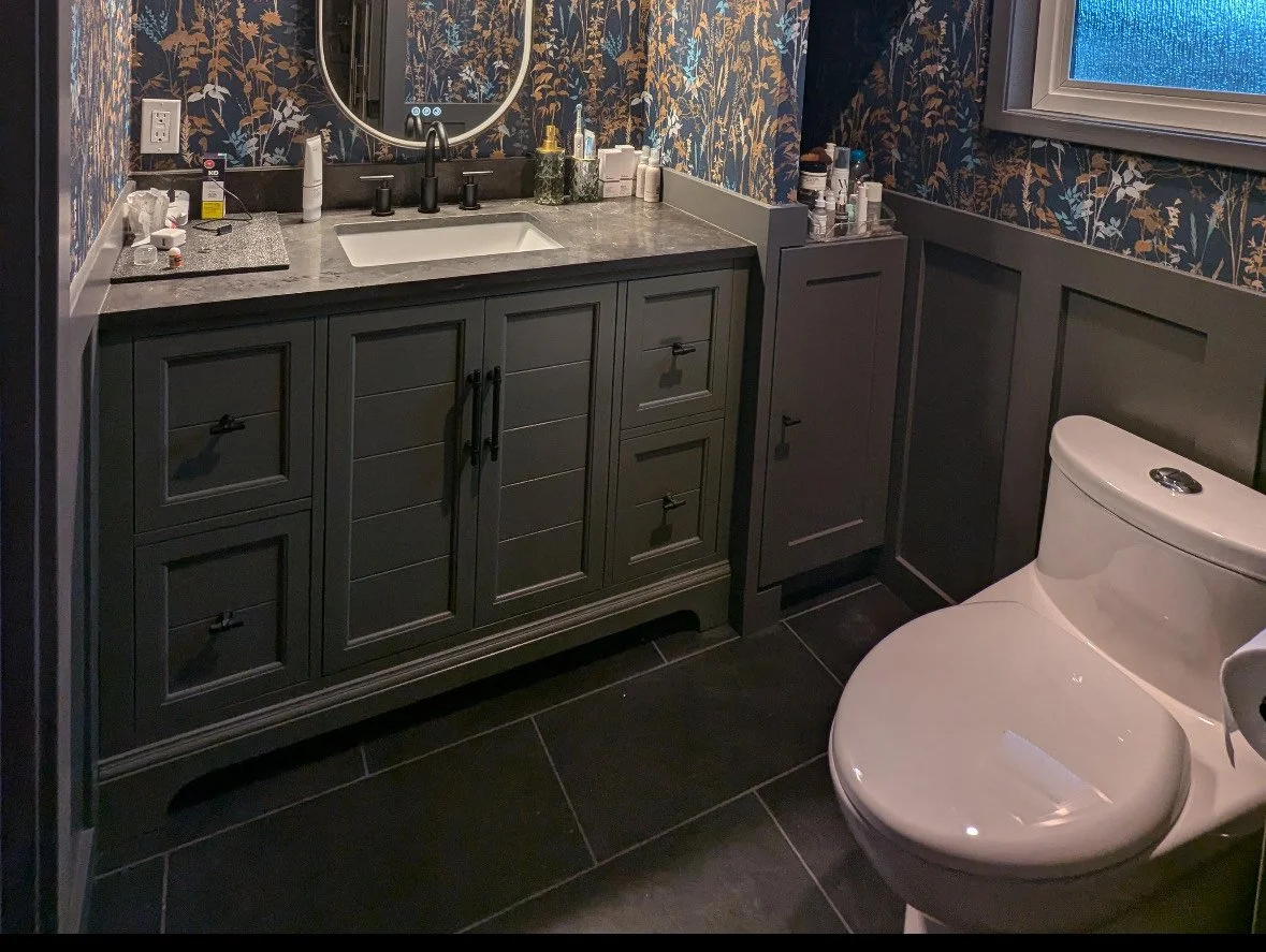 Bathroom with dark floral wallpaper, gray vanity with countertop, white sink, mirror, toilet, and window, with various toiletries on the counter and windowsill.