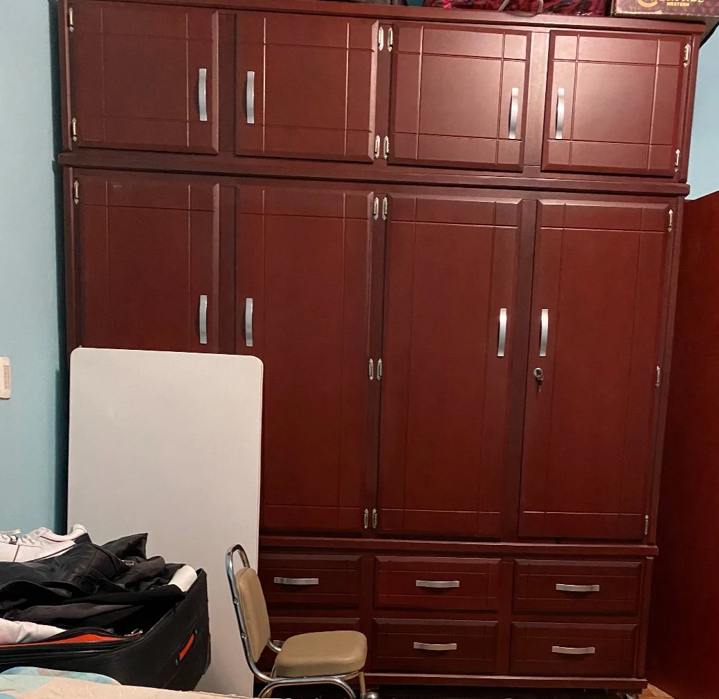 A large wooden wardrobe with multiple doors and drawers, and a small beige chair in front of it. There is an open suitcase with clothes next to the wardrobe and a white object leaning against it.