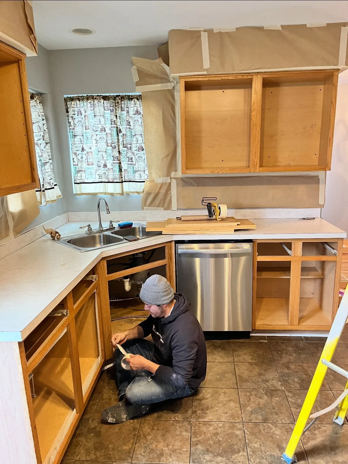 A person installing or repairing kitchen cabinets. The kitchen has a double sink, a small countertop dishwasher, and open cabinets with no doors. The person is sitting on the floor, wearing a gray beanie, black hoodie, and ripped jeans, working on the lower cabinets.