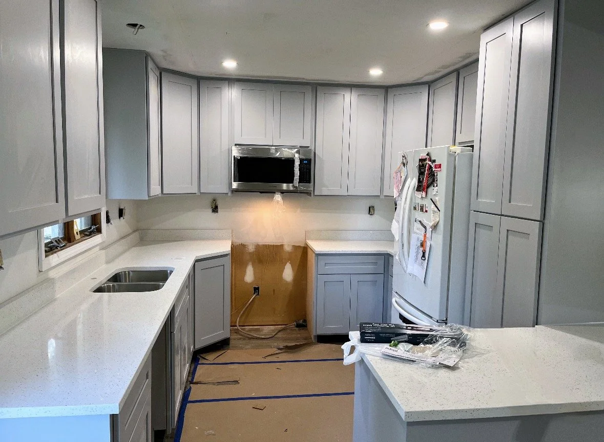 Kitchen with light gray cabinets, white countertops, a microwave above the stove, and a refrigerator. The area appears to be under renovation, with a cutout in the wall and construction materials on the floor and counter.