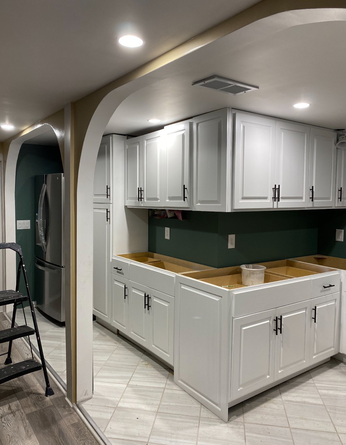 Kitchen under renovation with white cabinets, green walls, and unfinished countertop spaces. Refrigerator visible on the left, black ladder and construction items present.