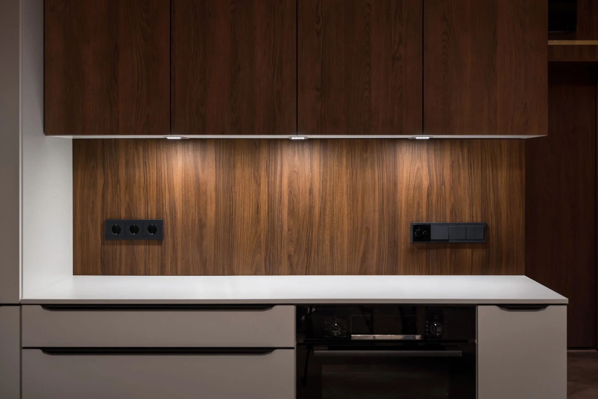 Modern kitchen with white countertops, wooden cabinets, and multiple electrical outlets on the wall.