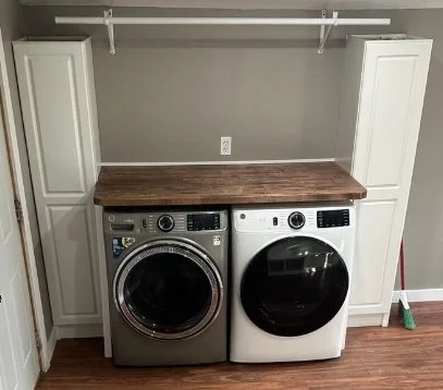 Laundry room with a front-loading washer and dryer beneath a wooden countertop, surrounded by white cabinets and a gray wall.