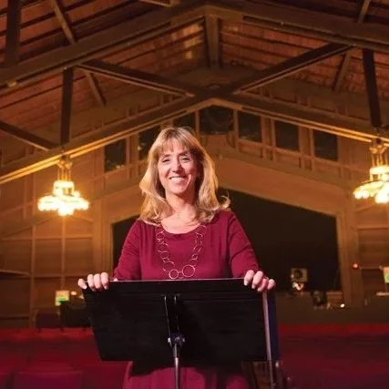 A woman with blonde hair wearing a maroon dress standing behind a music stand inside a wooden auditorium with warm lighting.