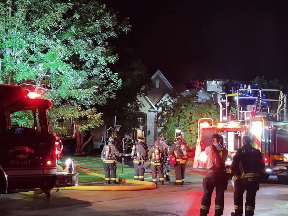 Nighttime scene of firefighters in gear responding to a fire on a residential street, with fire trucks and equipment, and a house partially visible in the background.